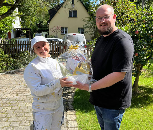 Fabian Sorgalla (rechts) überreicht Maler-Gesellin Jean zur bestandenen Gesellenprüfung draußen bei Sonnenschein einen Geschenkekorb von der Werkstück Raumgestaltung & Versorgungstechnik GmbH in Berlin. Fabian Sorgalla (rechts) überreicht Maler-Gesellin Jean zur bestandenen Gesellenprüfung draußen bei Sonnenschein einen Geschenkekorb von der Werkstück Raumgestaltung & Versorgungstechnik GmbH in Berlin.