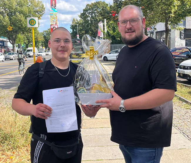 Fabian Sorgalla (rechts) überreicht Geselle Yanneck zur bestandenen Gesellenprüfung draußen bei Sonnenschein einen Geschenkekorb von der Werkstück Raumgestaltung & Versorgungstechnik GmbH in Berlin. Fabian Sorgalla (rechts) überreicht Geselle Yanneck zur bestandenen Gesellenprüfung draußen bei Sonnenschein einen Geschenkekorb von der Werkstück Raumgestaltung & Versorgungstechnik GmbH in Berlin.