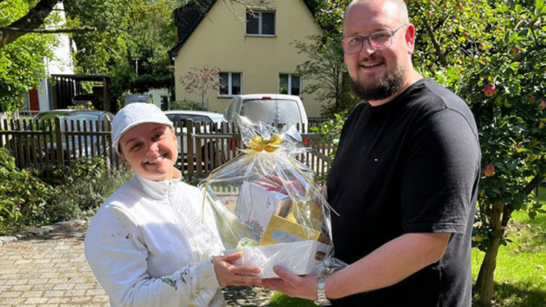 Fabian Sorgalla (rechts) überreicht Maler-Gesellin Jean zur bestandenen Gesellenprüfung draußen bei Sonnenschein einen Geschenkekorb von der Werkstück Raumgestaltung & Versorgungstechnik GmbH in Berlin. Fabian Sorgalla (rechts) überreicht Maler-Gesellin Jean zur bestandenen Gesellenprüfung draußen bei Sonnenschein einen Geschenkekorb von der Werkstück Raumgestaltung & Versorgungstechnik GmbH in Berlin.