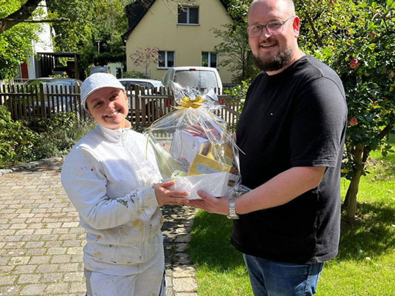 Fabian Sorgalla (rechts) überreicht Maler-Gesellin Jean zur bestandenen Gesellenprüfung draußen bei Sonnenschein einen Geschenkekorb von der Werkstück Raumgestaltung & Versorgungstechnik GmbH in Berlin. Fabian Sorgalla (rechts) überreicht Maler-Gesellin Jean zur bestandenen Gesellenprüfung draußen bei Sonnenschein einen Geschenkekorb von der Werkstück Raumgestaltung & Versorgungstechnik GmbH in Berlin.