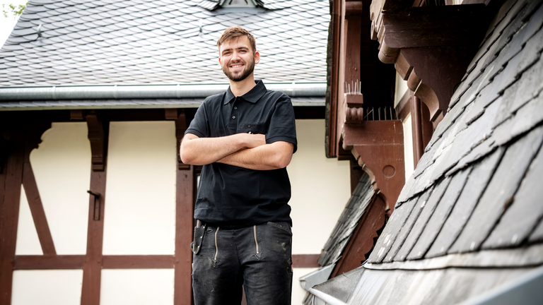 Auszubildender steht auf Dach und schaut mit verschränkten Armen zur Kamera Auszubildender steht auf Dach und schaut mit verschränkten Armen zur Kamera