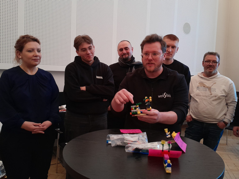 Ein Mann spricht zur umstehenden Gruppe an einem Tisch mit einem Segelboot aus bunten Legosteinen beim Teamtag vom Meisterbetrieb für Handwerk Werkstück Berlin zum Thema "Gute Kommunikation verbindet" in Berlin-Wannsee