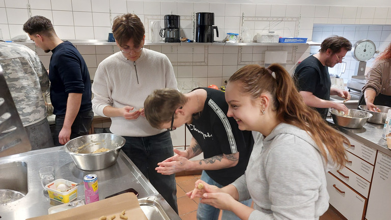 Drei junge Auszubildende stehen um einem Tisch mit einem Backblech und Schüsseln, unterhalten sich und kneten Plätzchenteig in der Küche der Gemeinde der Baptisten in Berlin-Schönberg beim 4. Azubitag der Auszubildenden von Werkstück Berlin mit Unterstützung von Immanuel Dienstleistungen